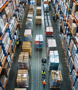 High-angle view of workers managing pallets of boxed goods in a large warehouse aisle lined with tall storage racks.
