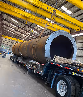 Large cylindrical metal pipe being lifted by chains inside an industrial warehouse onto a flatbed truck.